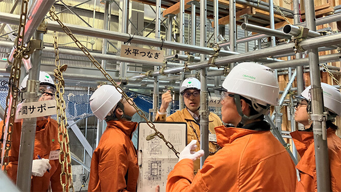 足場が組まれた作業環境で、図面を確認しながら複数名が説明を受けている様子